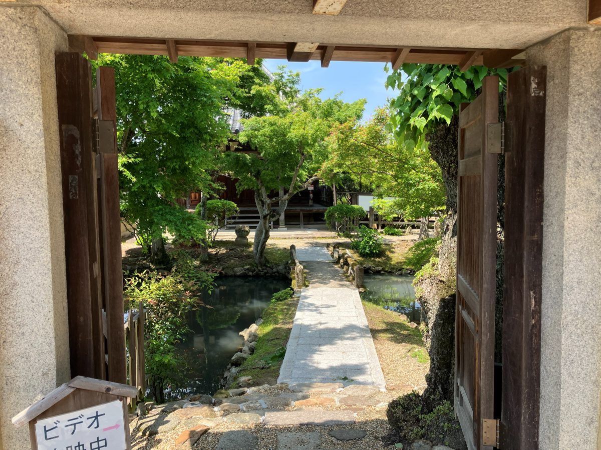 An entrance to the garden of Shin-Yakushiji, with a pond and stone bridge beyond the gate and a video sign